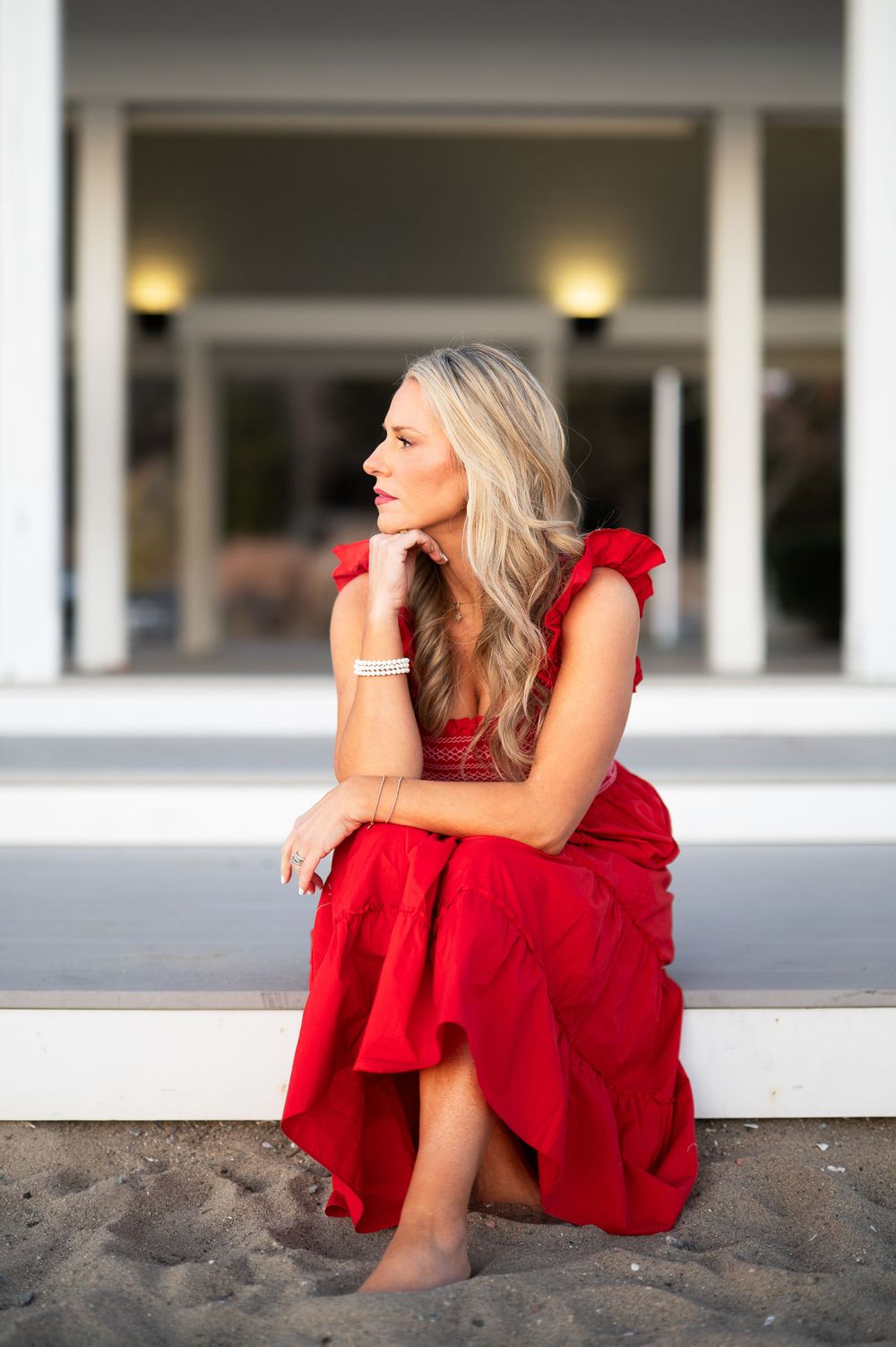 Woman in flowing red dress sits elegantly on beach wall at sunset with soft lighting and architecture in background.