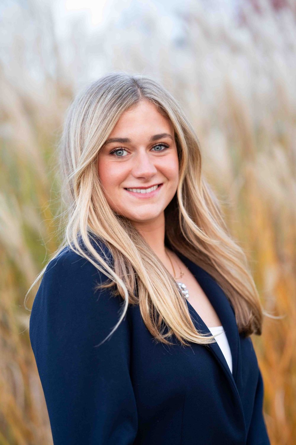 Professional headshot portrait of someone in a navy blazer and white top smiling outdoors with autumn grass in background.