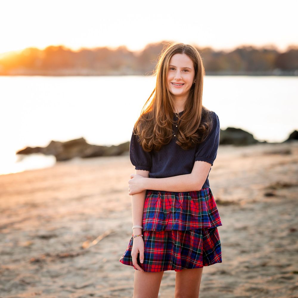 Person in plaid dress and black top poses on sandy beach at sunset with ocean waves in background.