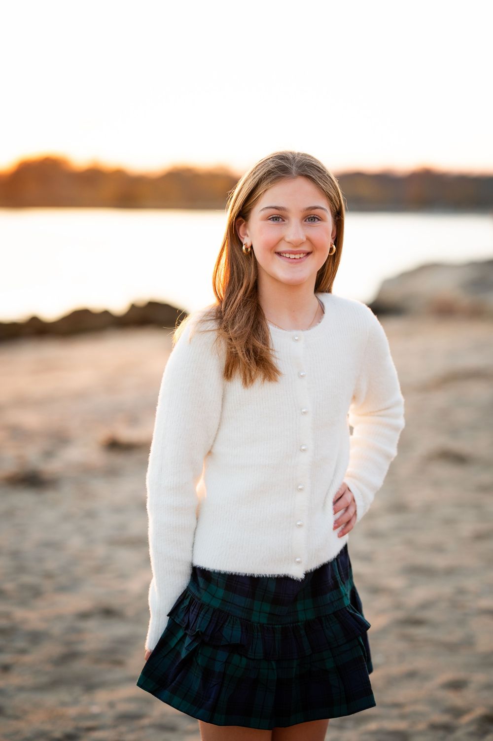 A person wearing a white sweater and plaid skirt poses on a beach at sunset with water in the background.