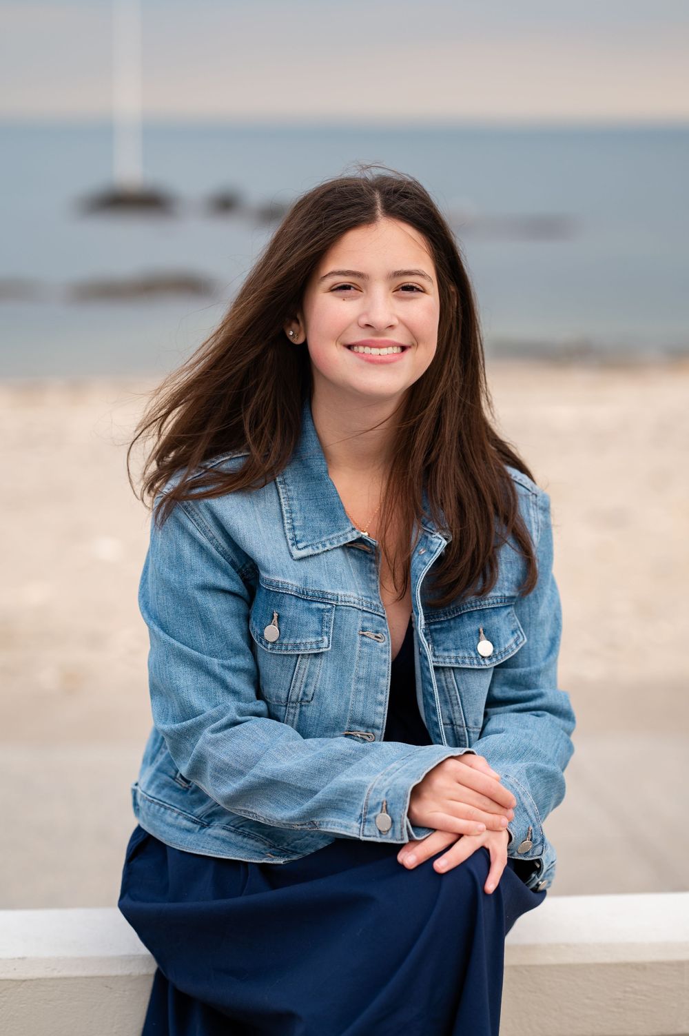 Person in denim jacket poses casually on beach with soft ocean waves and rocks in background.