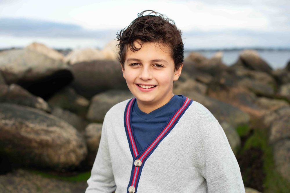 A young person in a gray cardigan smiles while sitting near rocky coastline on an overcast day.