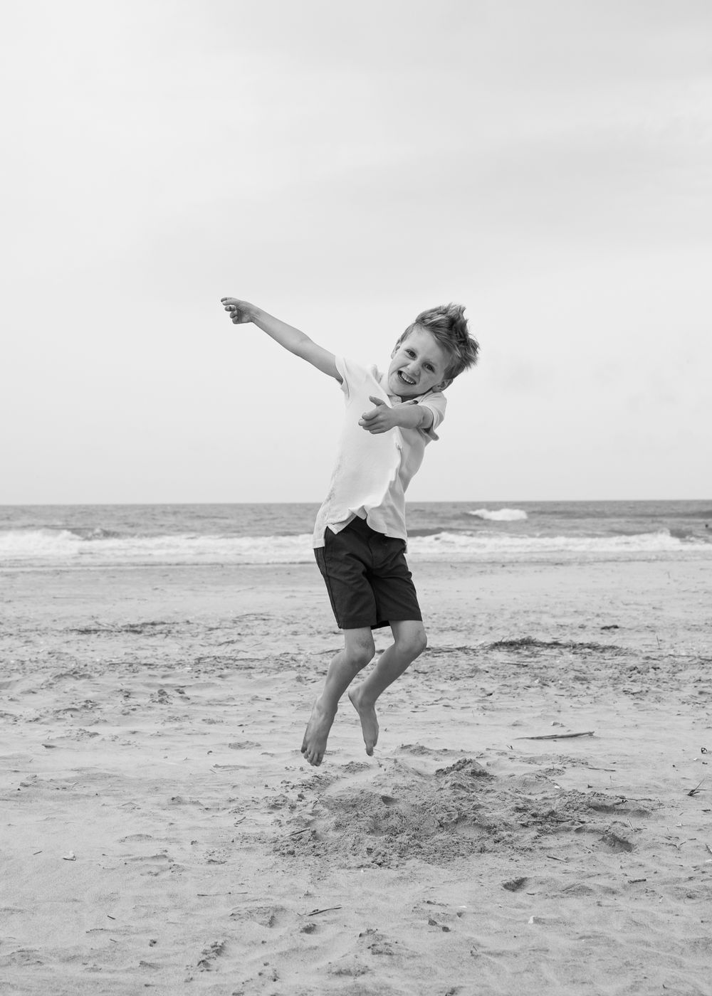 A child joyfully leaps and plays on a sandy beach with the ocean waves visible in the black and white background.