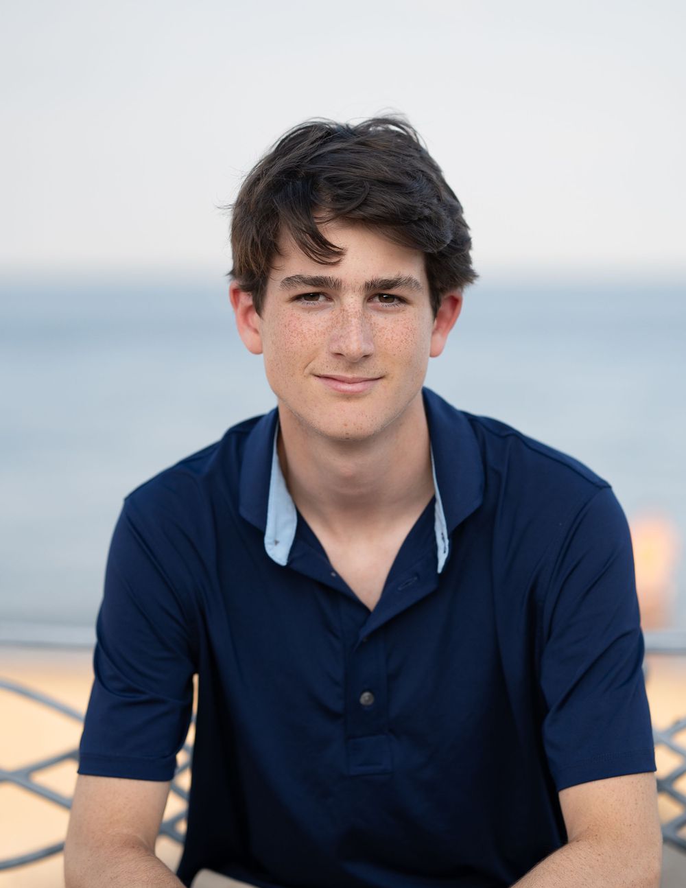 Senior high school boy wearing a navy polo shirt poses outdoors against a soft blurred waterfront background at dusk.