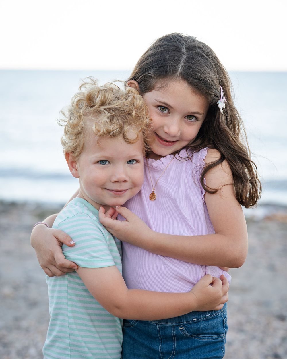 Two children hugging and smiling together at the beach with ocean in background during sunset.