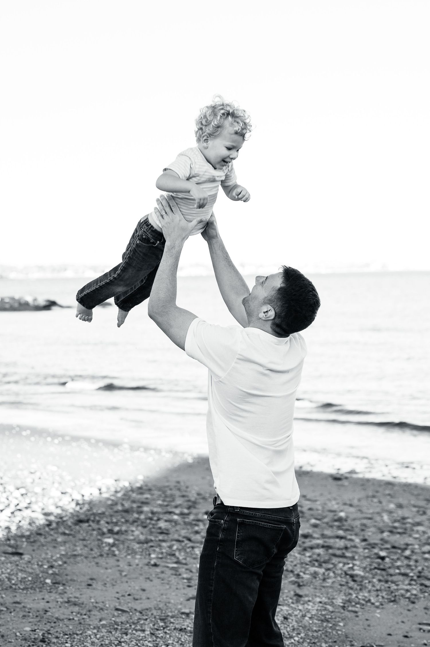 A black and white photograph of a parent lifting a joyful toddler high in the air at the beach.