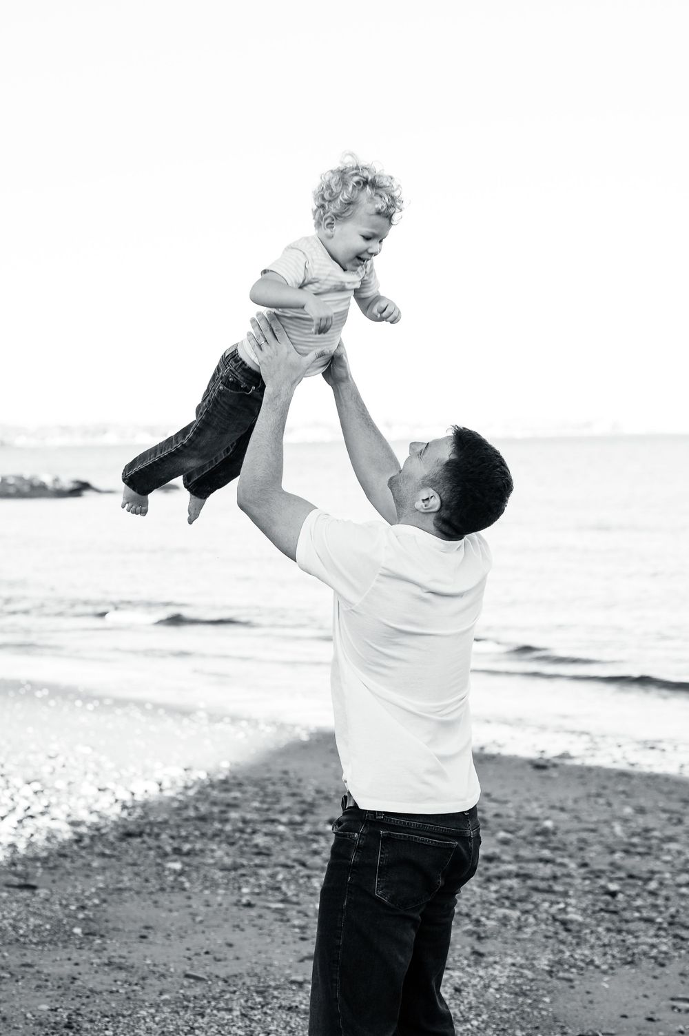 A black and white photograph of a parent lifting a joyful toddler high in the air at the beach.