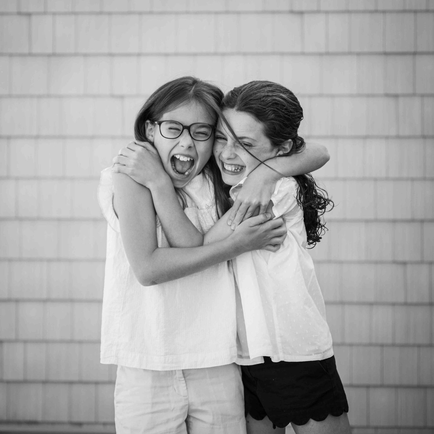 Two friends in white tops laughing and hugging joyfully against a light background in black and white photography.