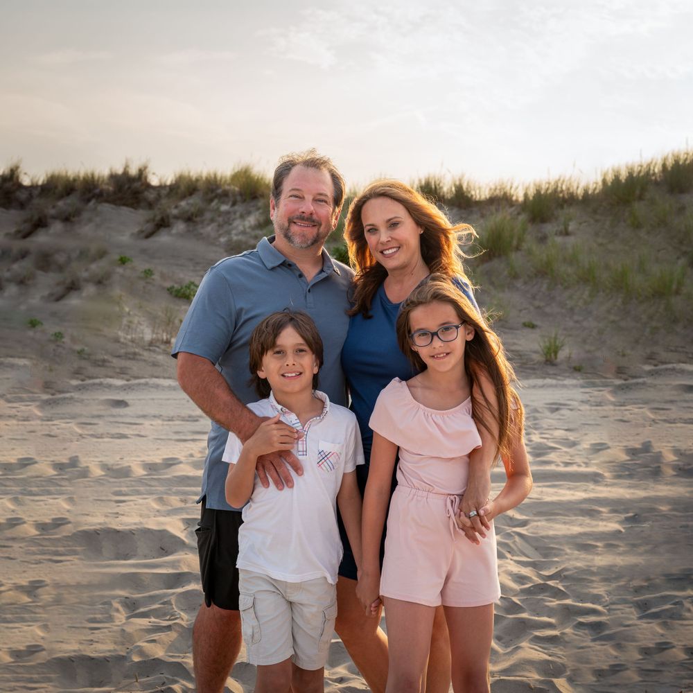 A family poses together on sandy beach dunes at sunset during a casual beach photoshoot.