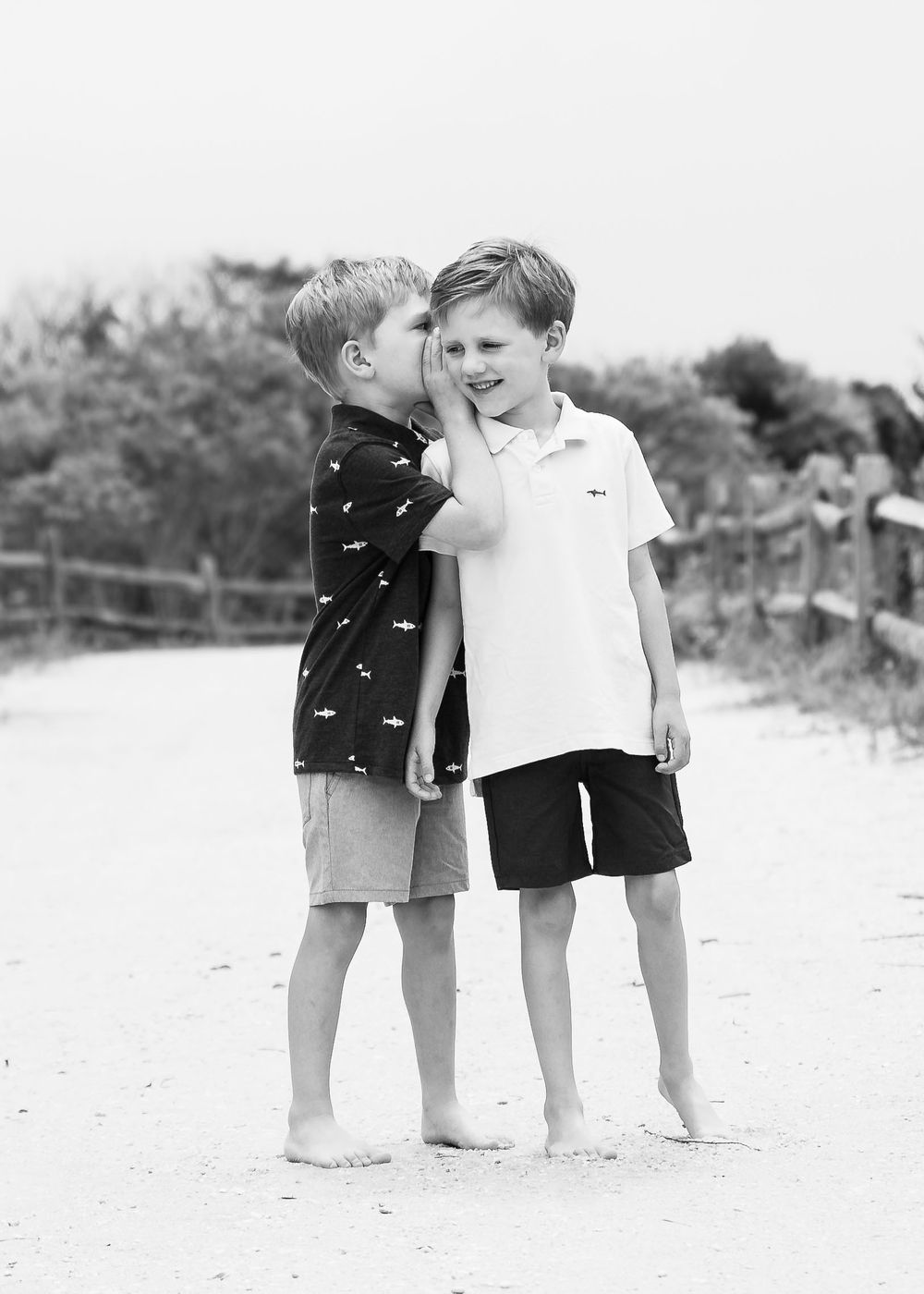 Black and white photo showing two young boys sharing a secret on a sandy beach path.