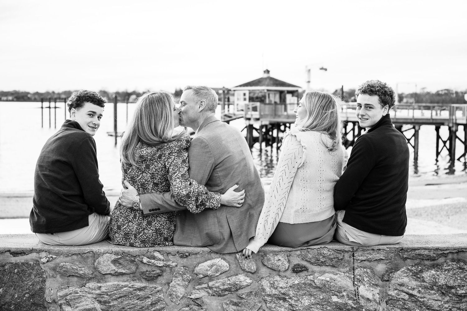 A group of friends sit together on a stone wall overlooking a pier on a sunny day, captured in black and white.