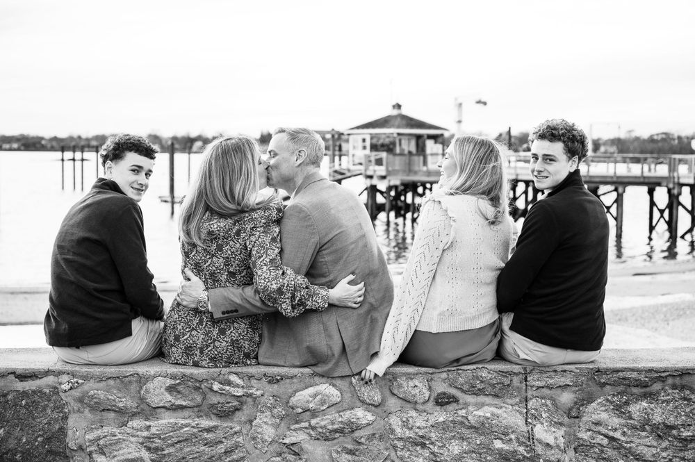 A group of friends sit together on a stone wall overlooking a pier on a sunny day, captured in black and white.