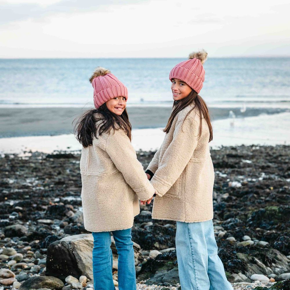 Two sisters in matching pink beanies and beige coats hold hands while enjoying a rocky beach on an overcast day.
