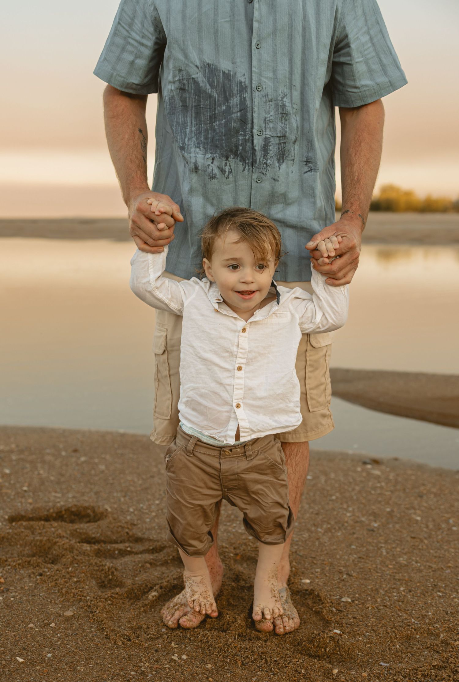 From ankle to neck, a person wearing a blue shirt and khaki shorts stands next to a child in white shirt and khaki shorts.