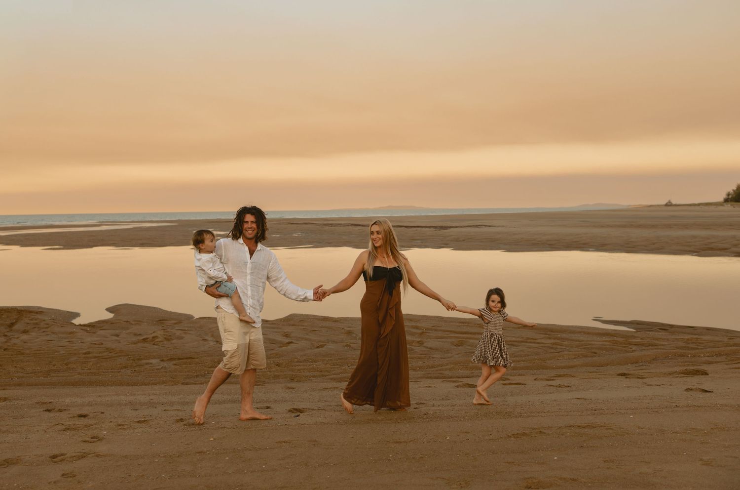 Three people in flowy outfits dance gracefully on a sandy beach at sunset creating a dreamy coastal scene.