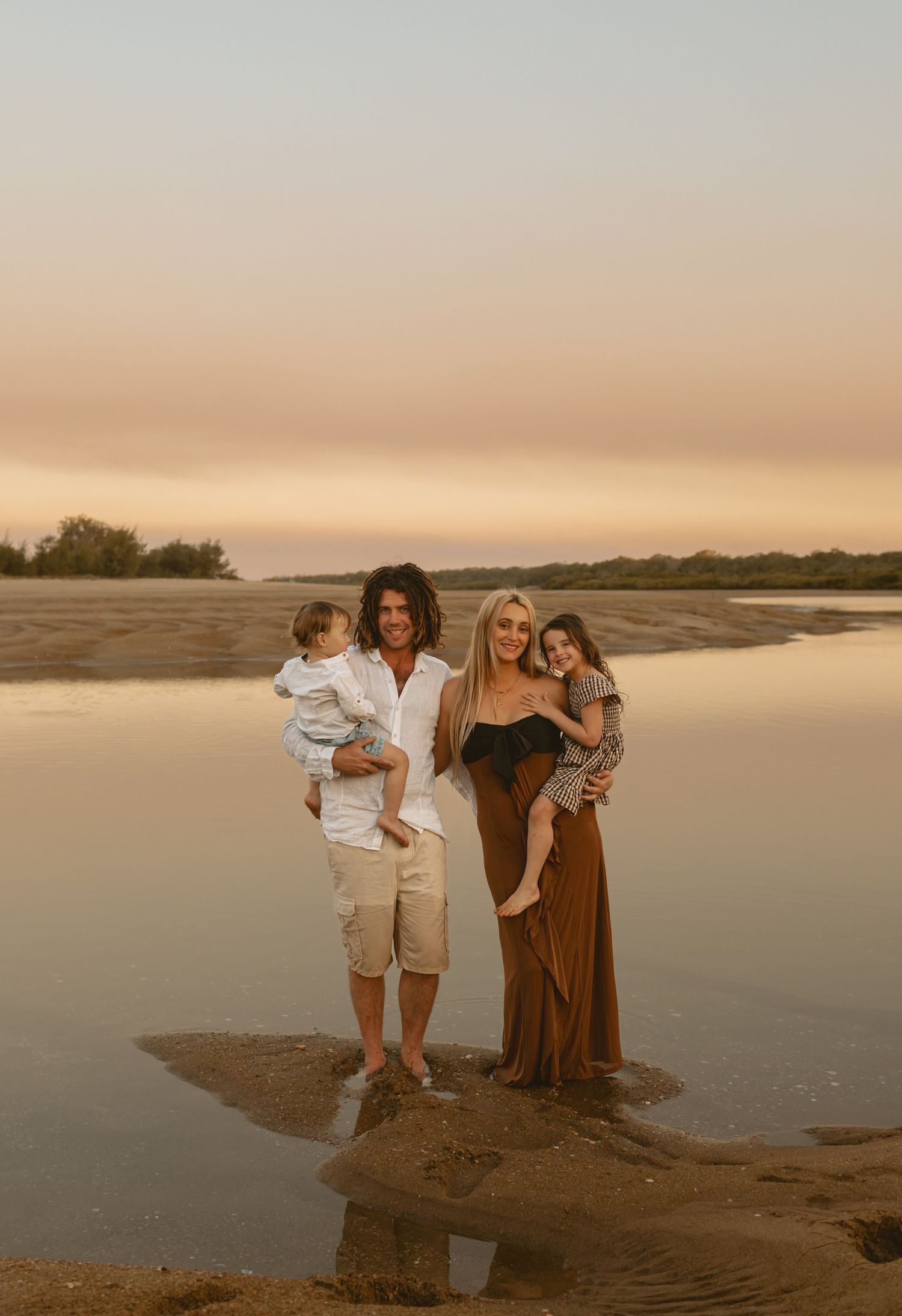 Family group poses together on sandy beach at sunset with calm water and warm lighting creating a serene atmosphere.