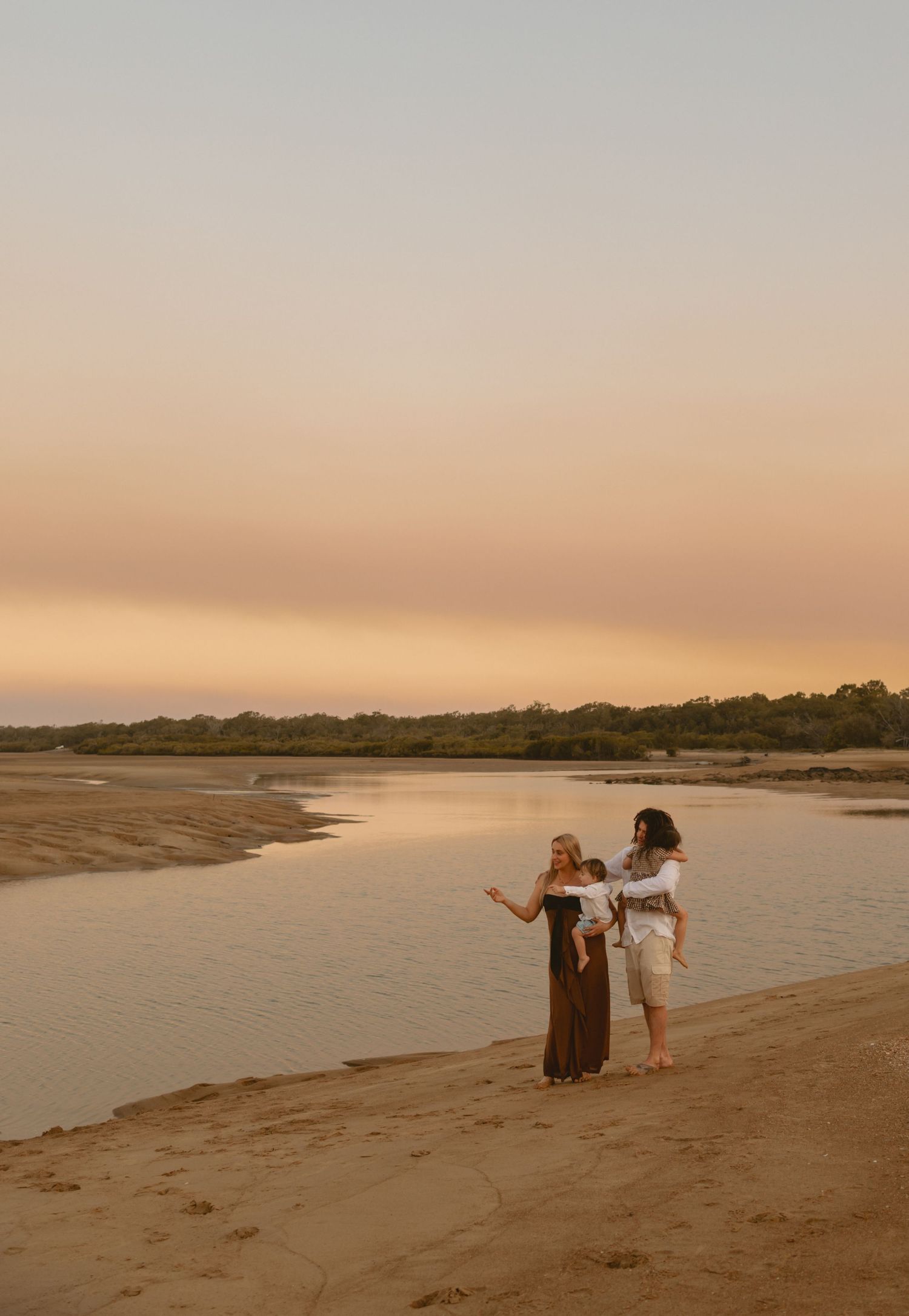 People walk along a sandy beach at sunset with warm golden light reflecting off the calm waters of the shoreline.