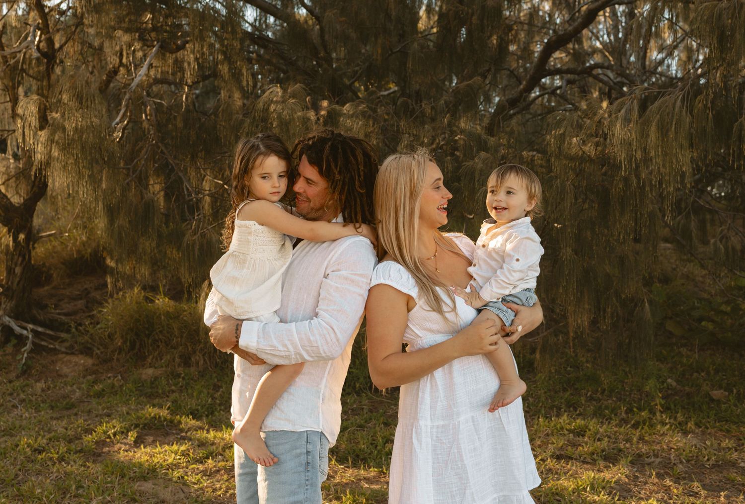 A loving family in white outfits embraces together during golden hour in a wooded outdoor setting.