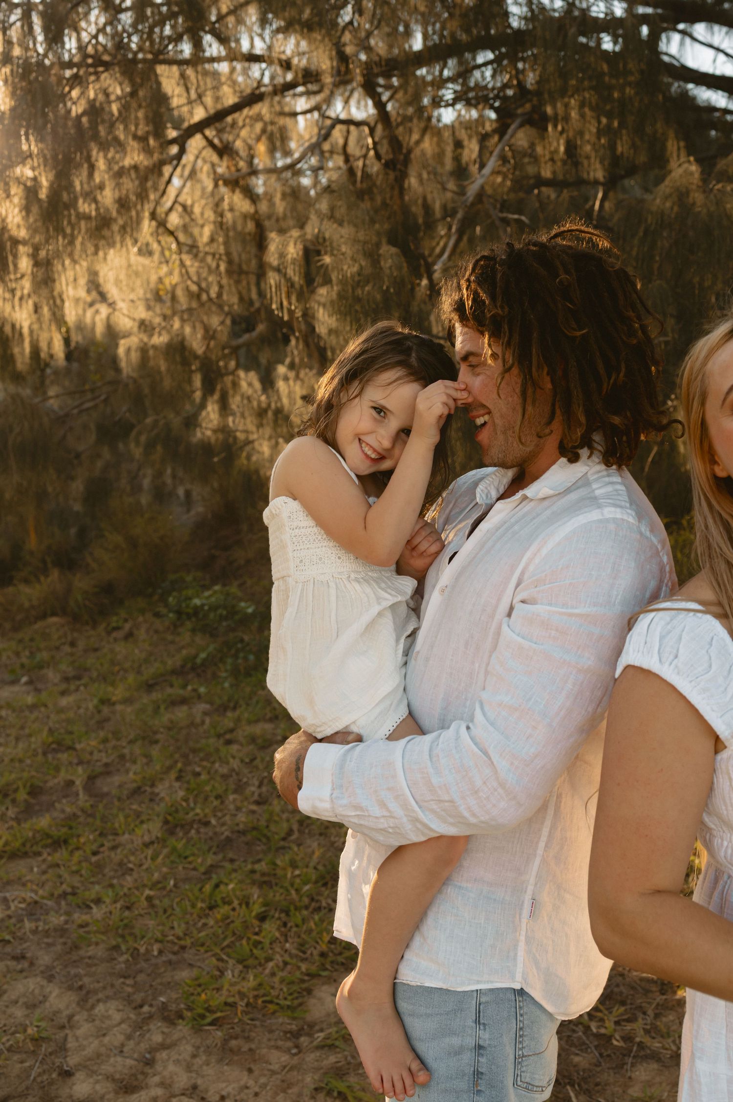 Couple in white clothing embraces outdoors at golden hour in a wooded setting.