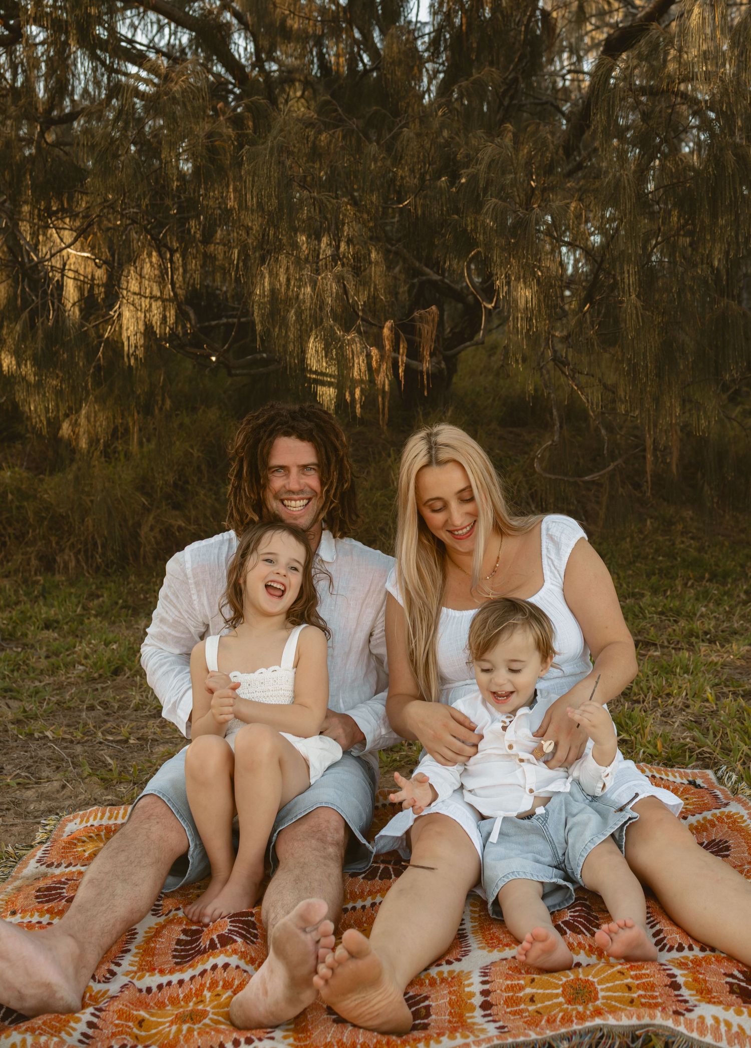 A family shares joyful moments together while relaxing on an orange blanket in a wooded setting during sunset.