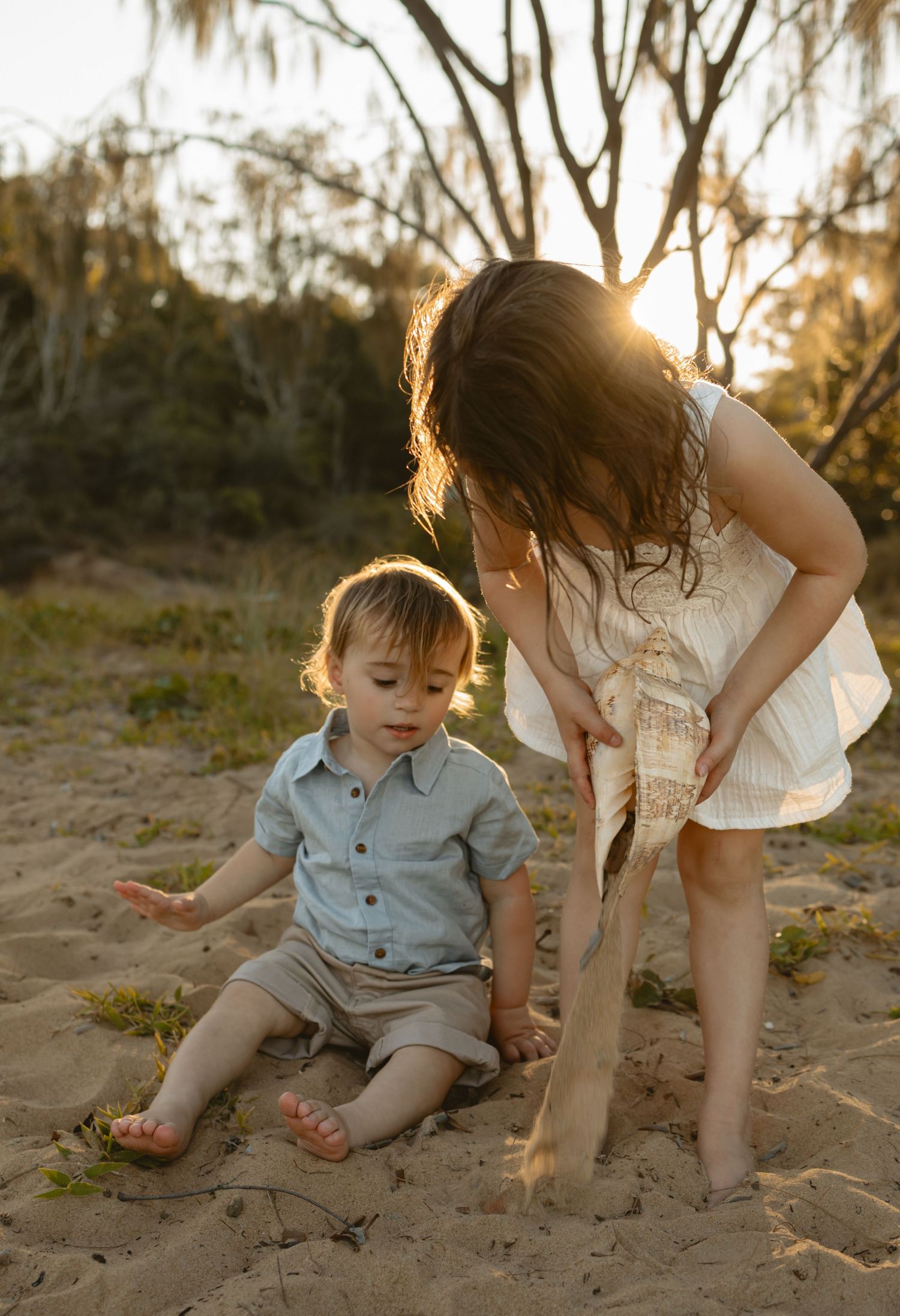 Small child wearing blue plays in soft beach sand at sunset with sunlit trees in background.