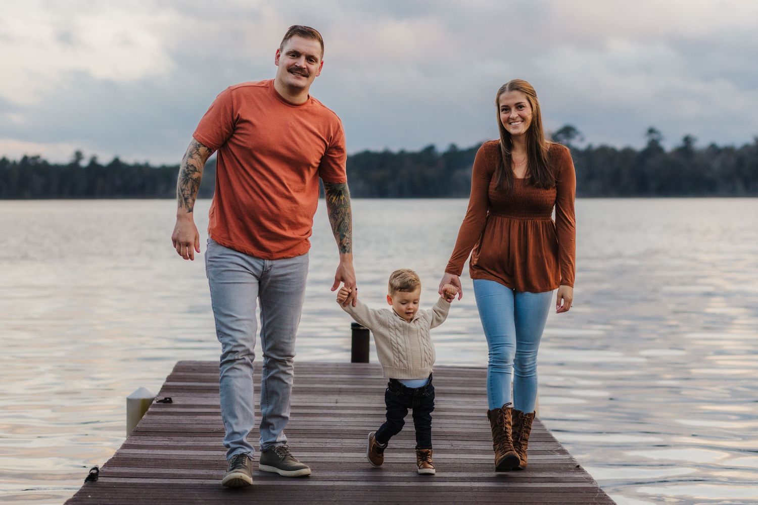 Family walks together on wooden dock overlooking peaceful lake at sunset.
