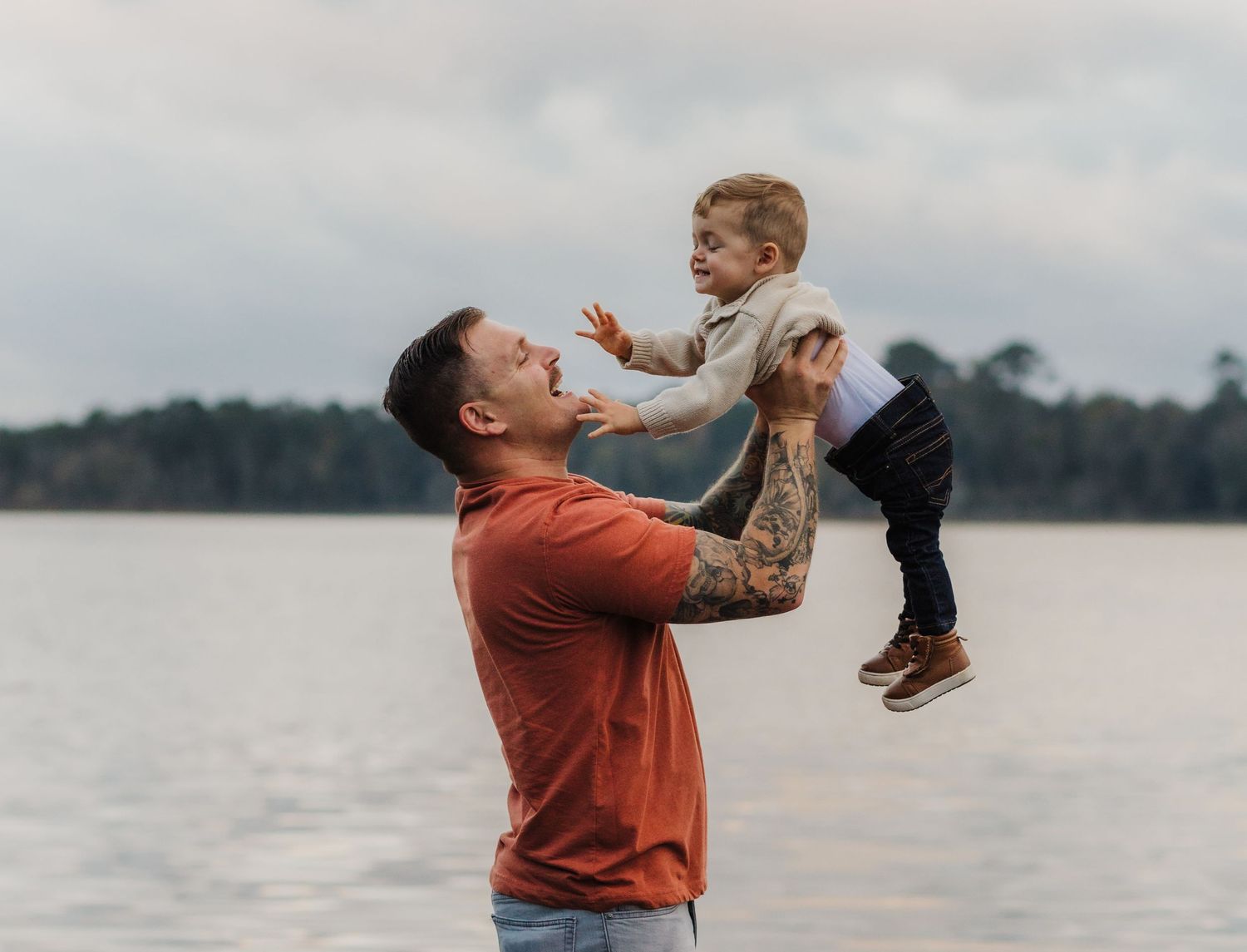 Joyful moment captured as parent lifts child high in the air by lakeside.