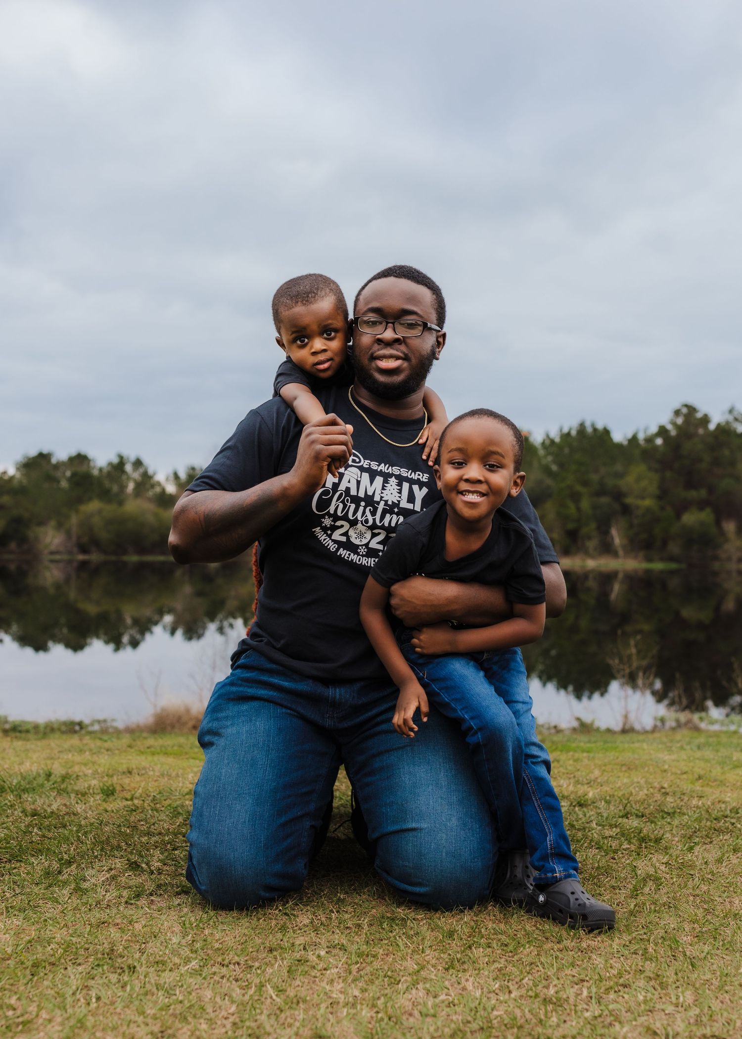Family group poses together on grass near a serene lake on an overcast day.