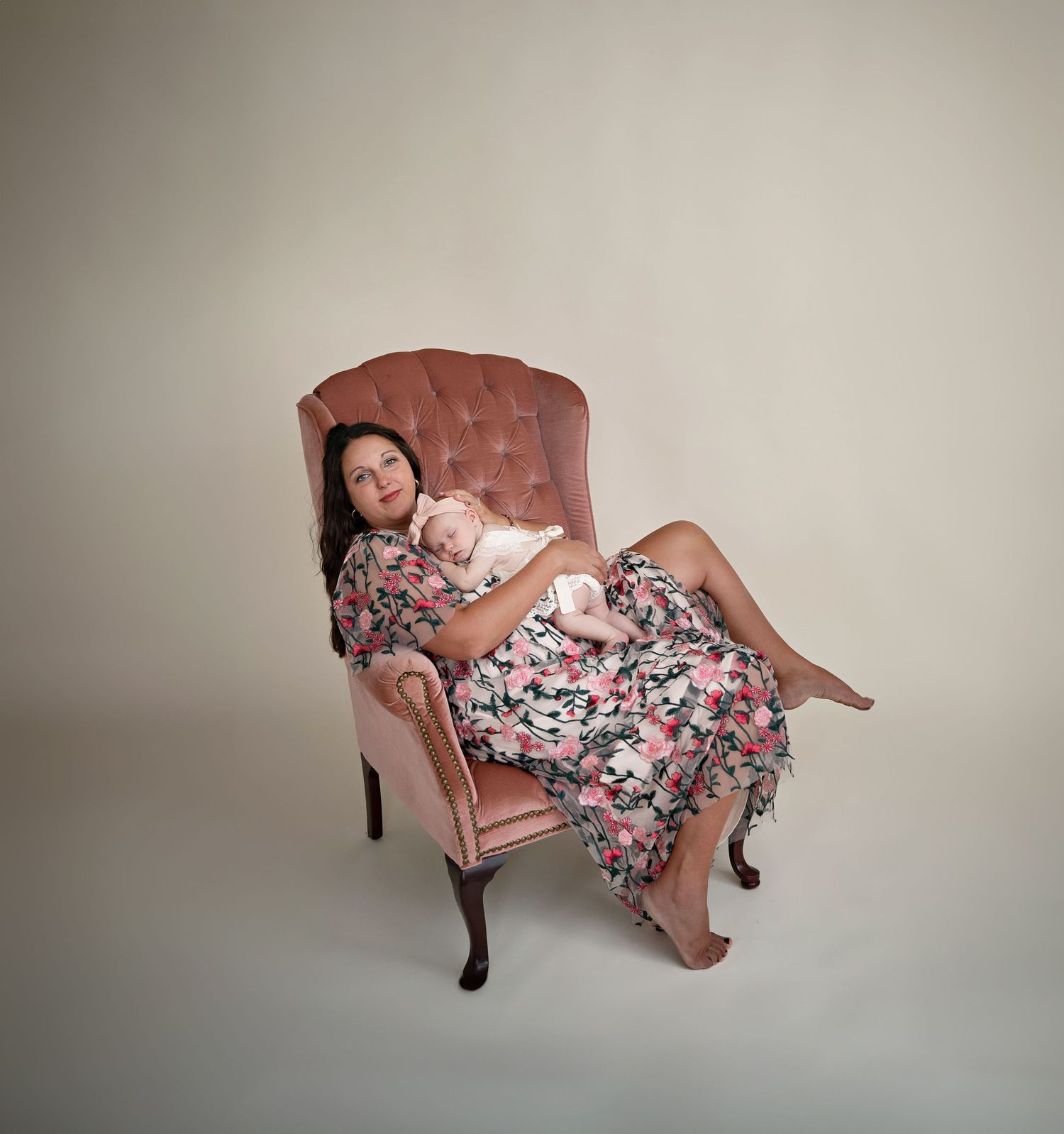 mother in a floral gown in a pink wingback chair holding a baby girl in a lace romper