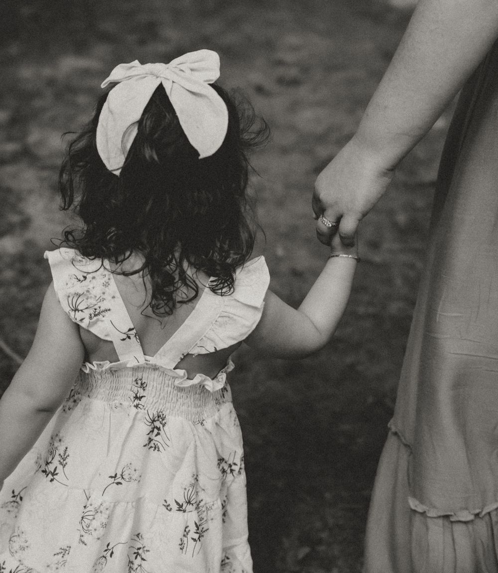 A black and white image of a small child in a floral dress with a bow holding an adult's hand. Colorado Springs Photographer.