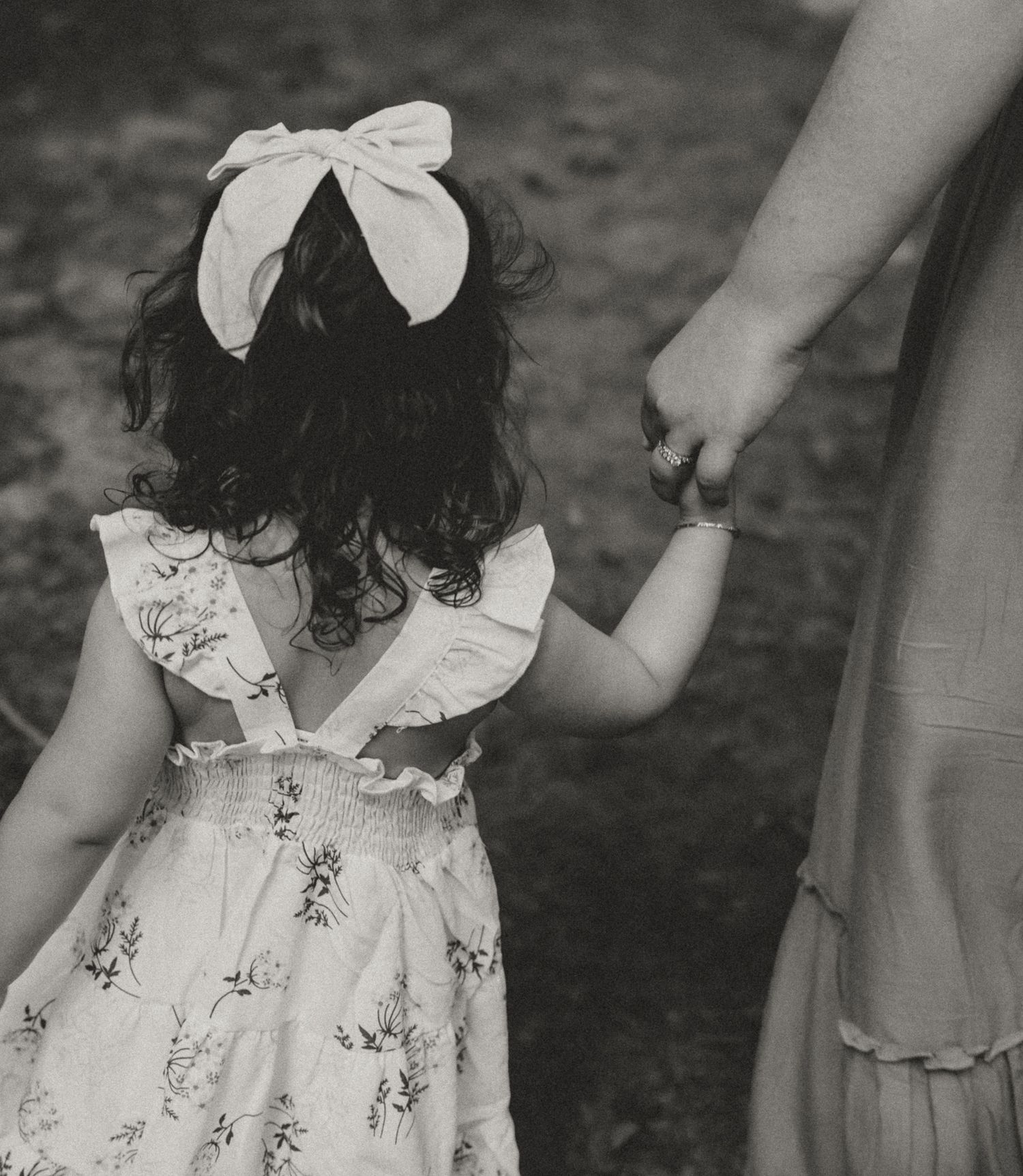 A black and white image of a small child in a floral dress with a bow holding an adult's hand. Colorado Springs Photographer.