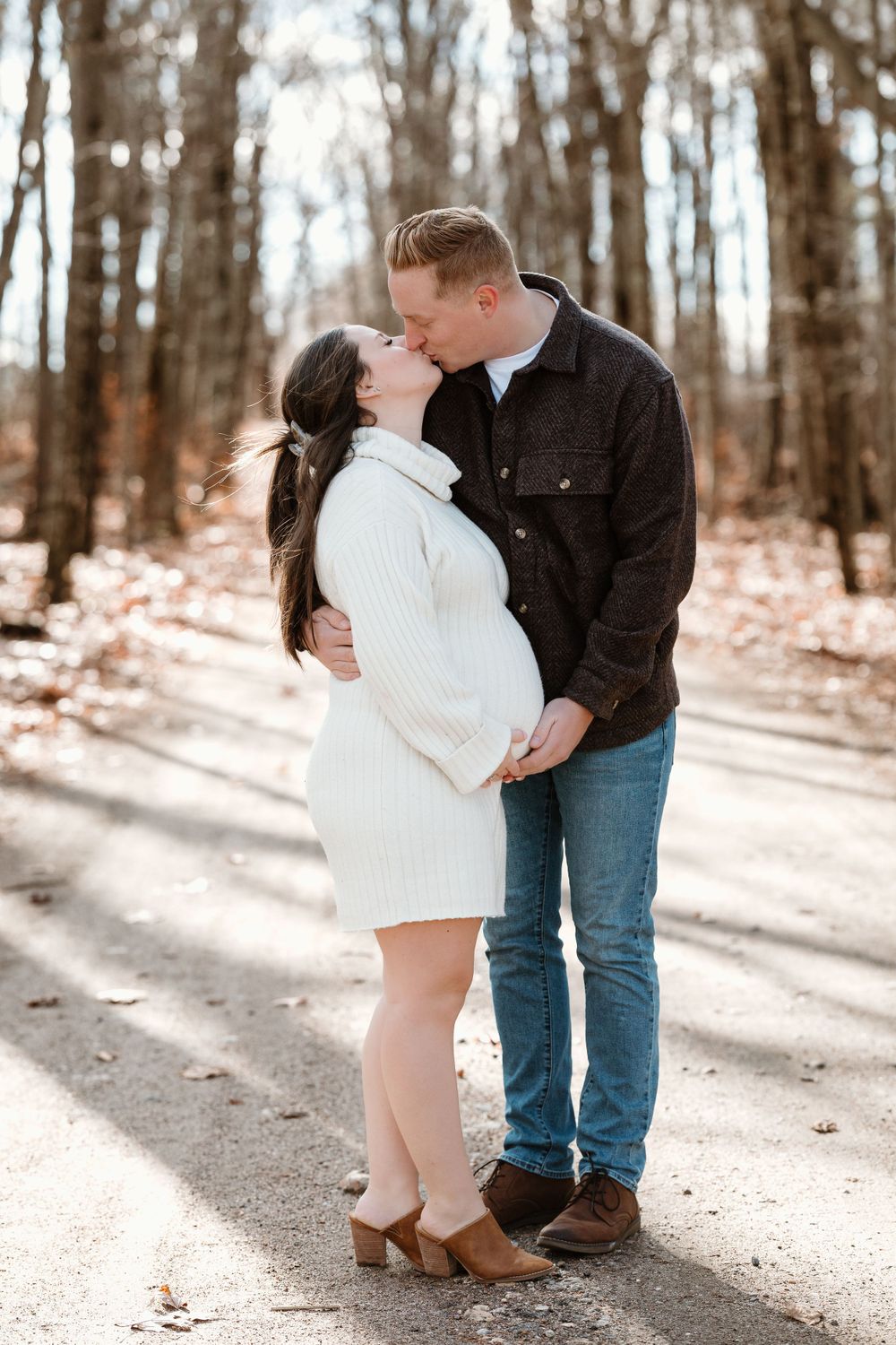 Winter maternity photo of a couple sharing a kiss on a snowy forest path.