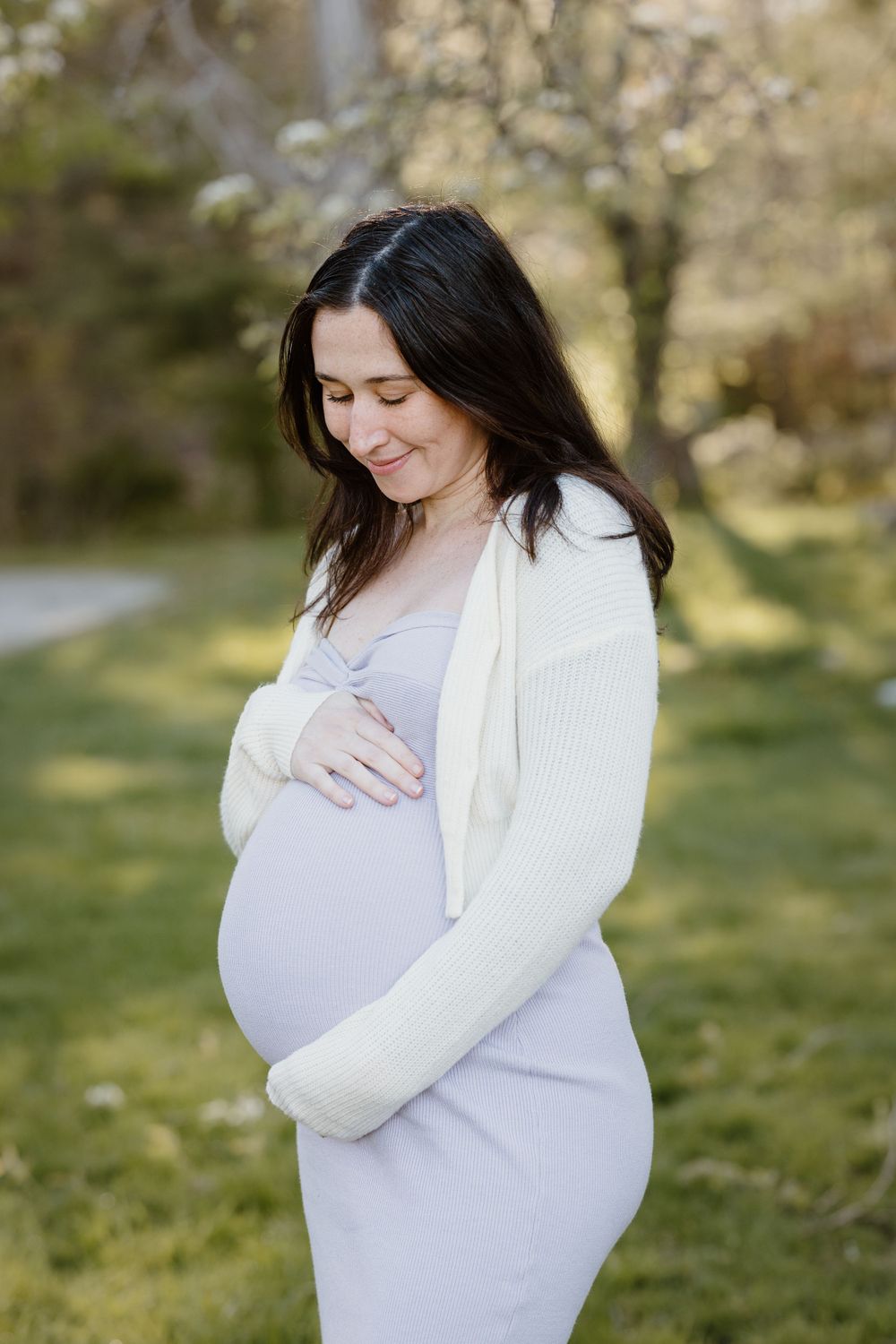An expectant mother in a white dress cradles her baby bump in a spring garden setting.