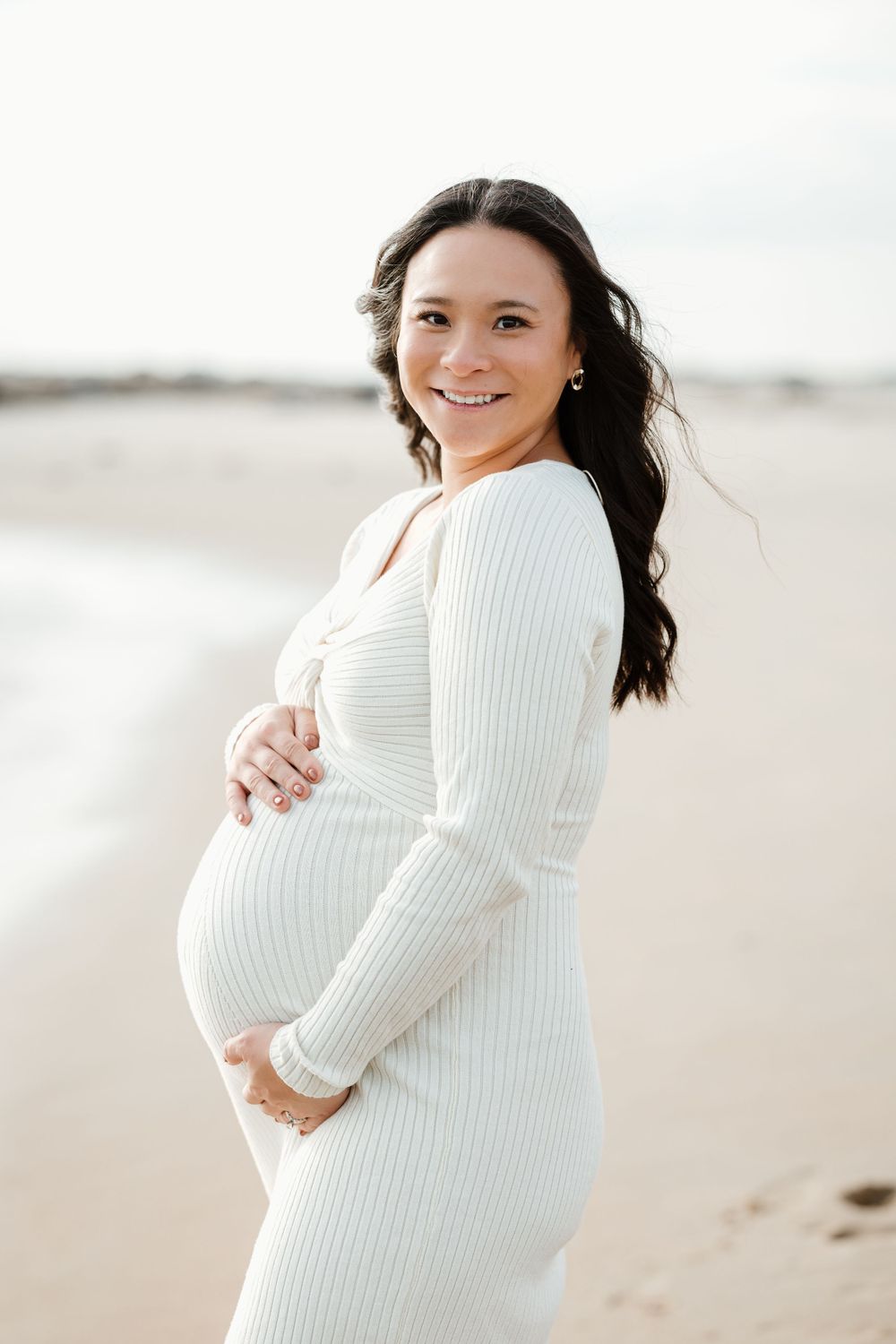Maternity portrait of woman in white ribbed dress on beach.