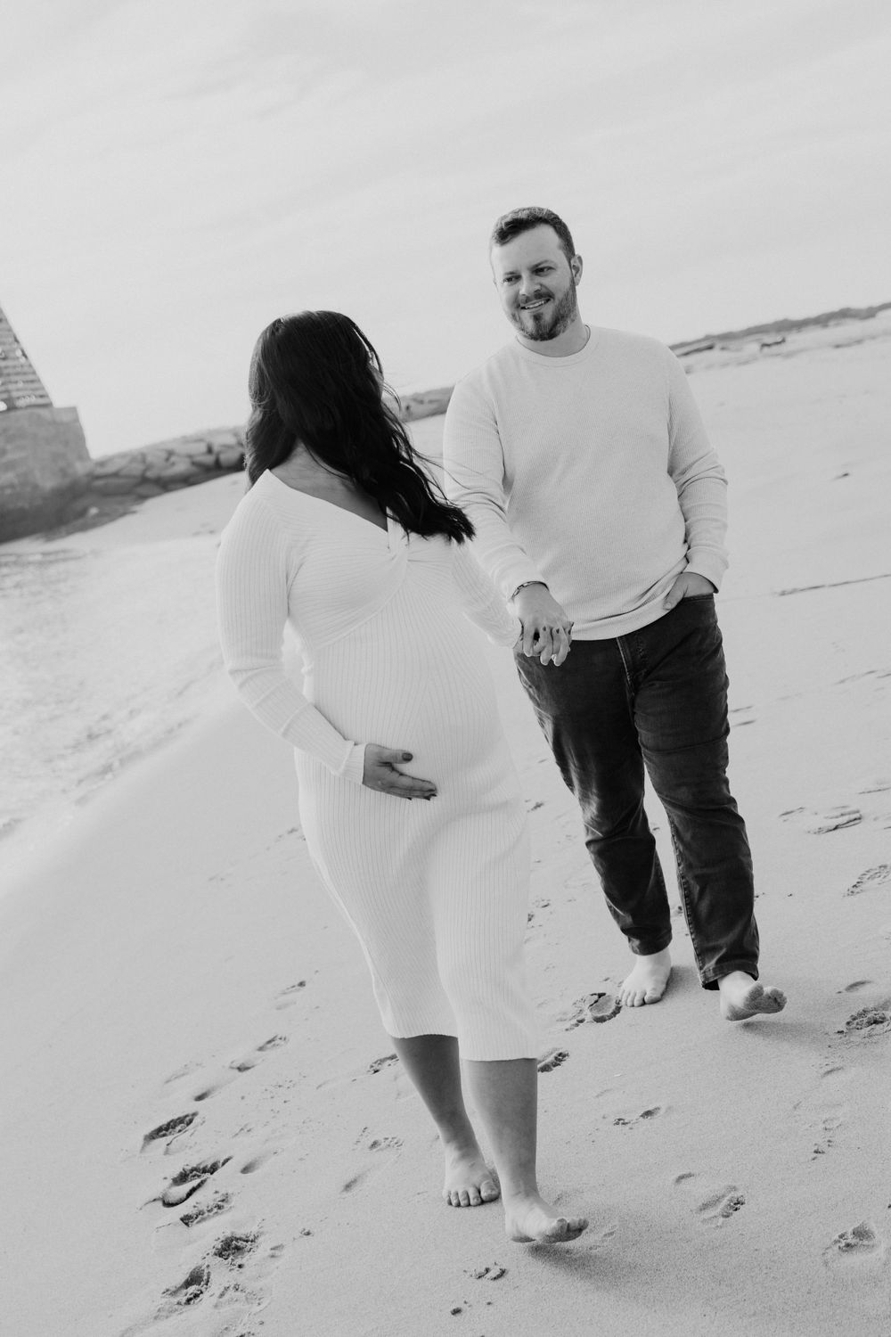 Black and white maternity photo of couple walking barefoot on beach.