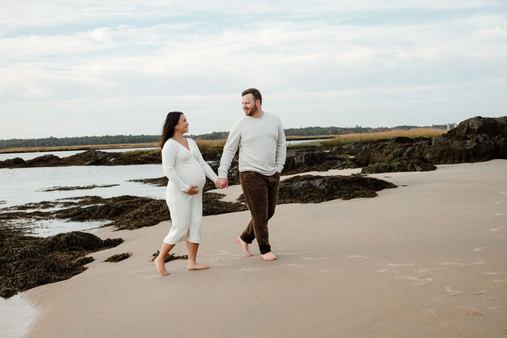 Expecting couple walking along rocky coastline in casual white attire.
