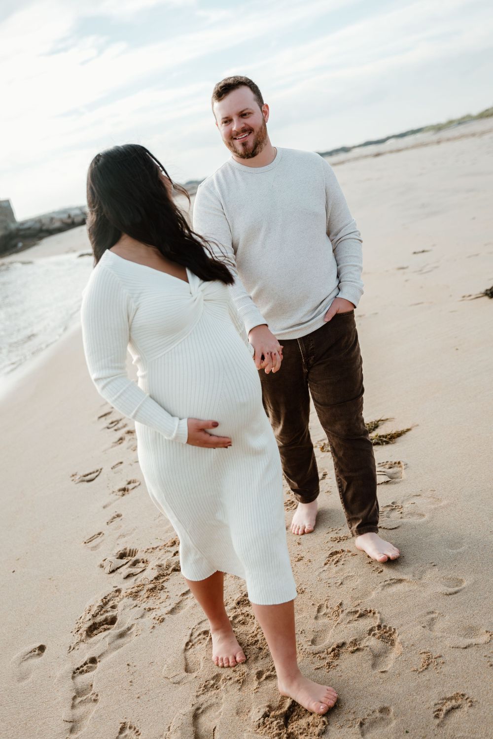 Expecting couple in matching white sweaters walking barefoot on sandy beach.