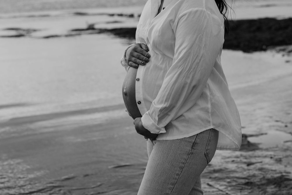 A black and white photo of someone embracing their pregnant belly at the beach.