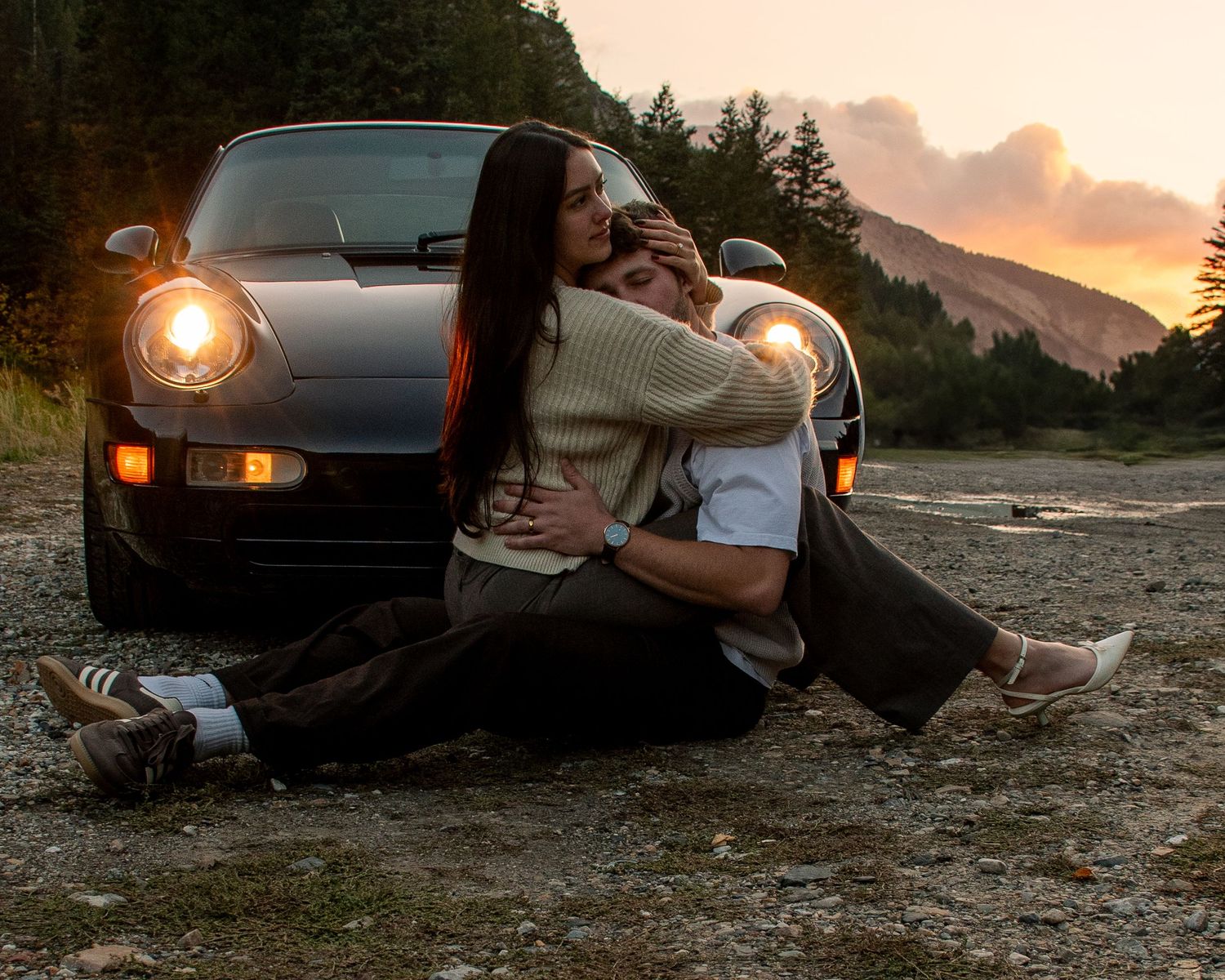 A boy and girl hug in front of a luxury car with a  stunning Utah sunset and mountain in the background.