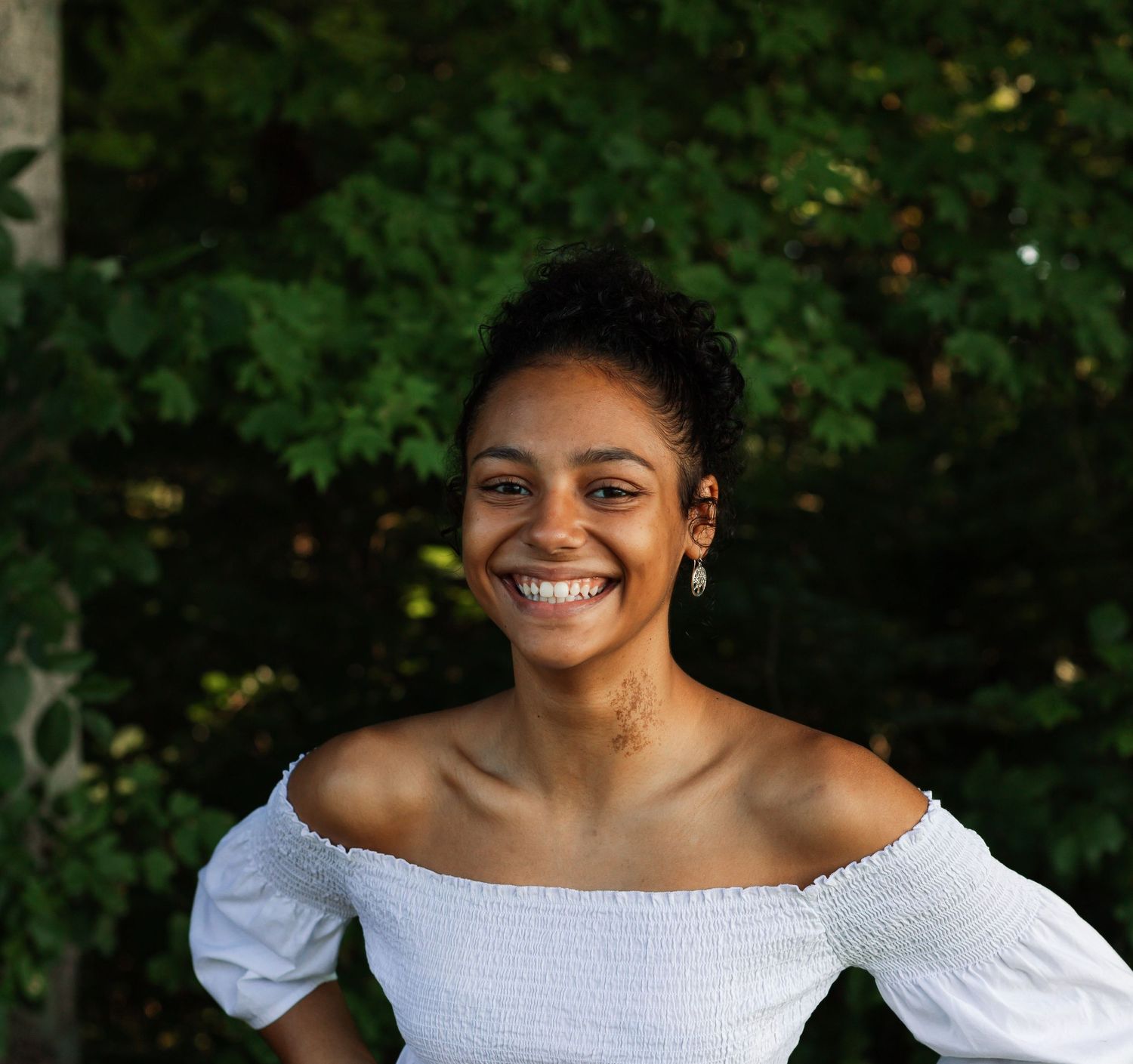 Portrait session outdoors featuring bright smile and white off-shoulder top against green foliage.