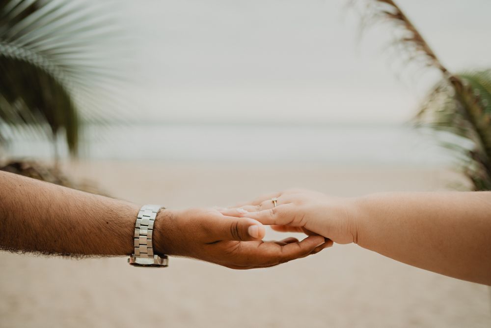 Bride resting on groom’s chest, ocean and sand, palm trees