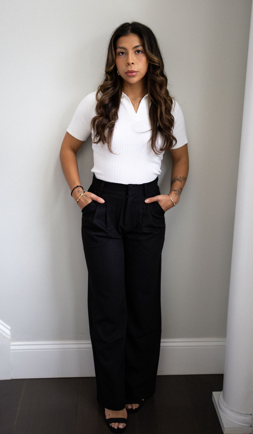 Young woman with long wavy hair standing against white wall in white ribbed top and wide-leg black trousers.