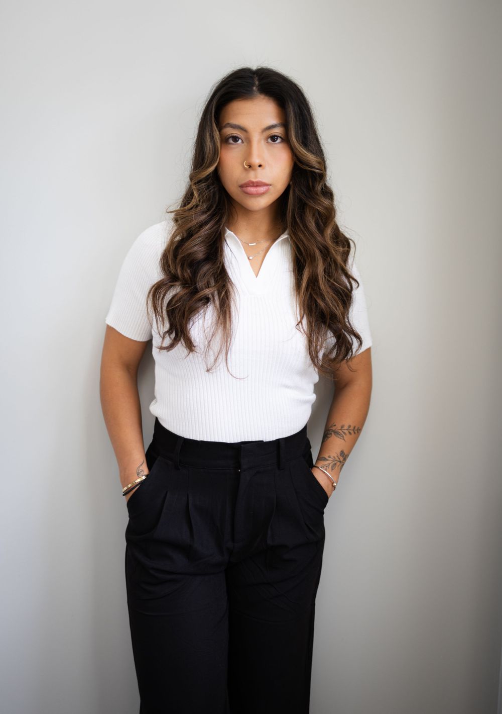 Young female aesthetic professional in white top and black pants posing confidently against a neutral wall.