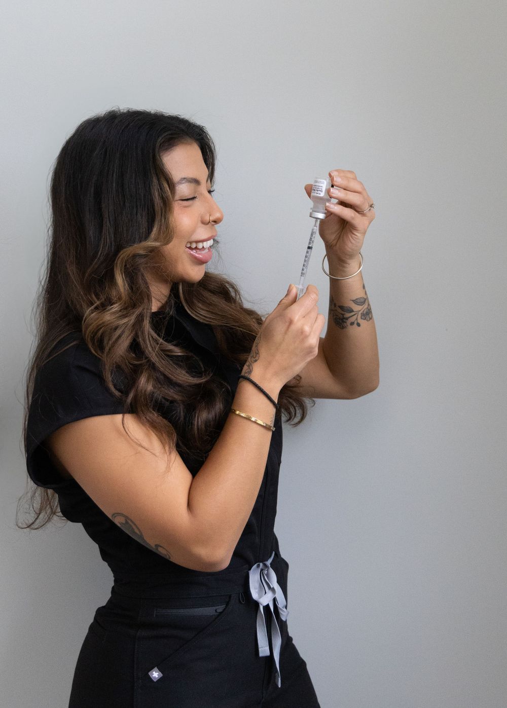 Female aesthetic injector in black scrubs laughing while preparing a syringe with injectable filler.