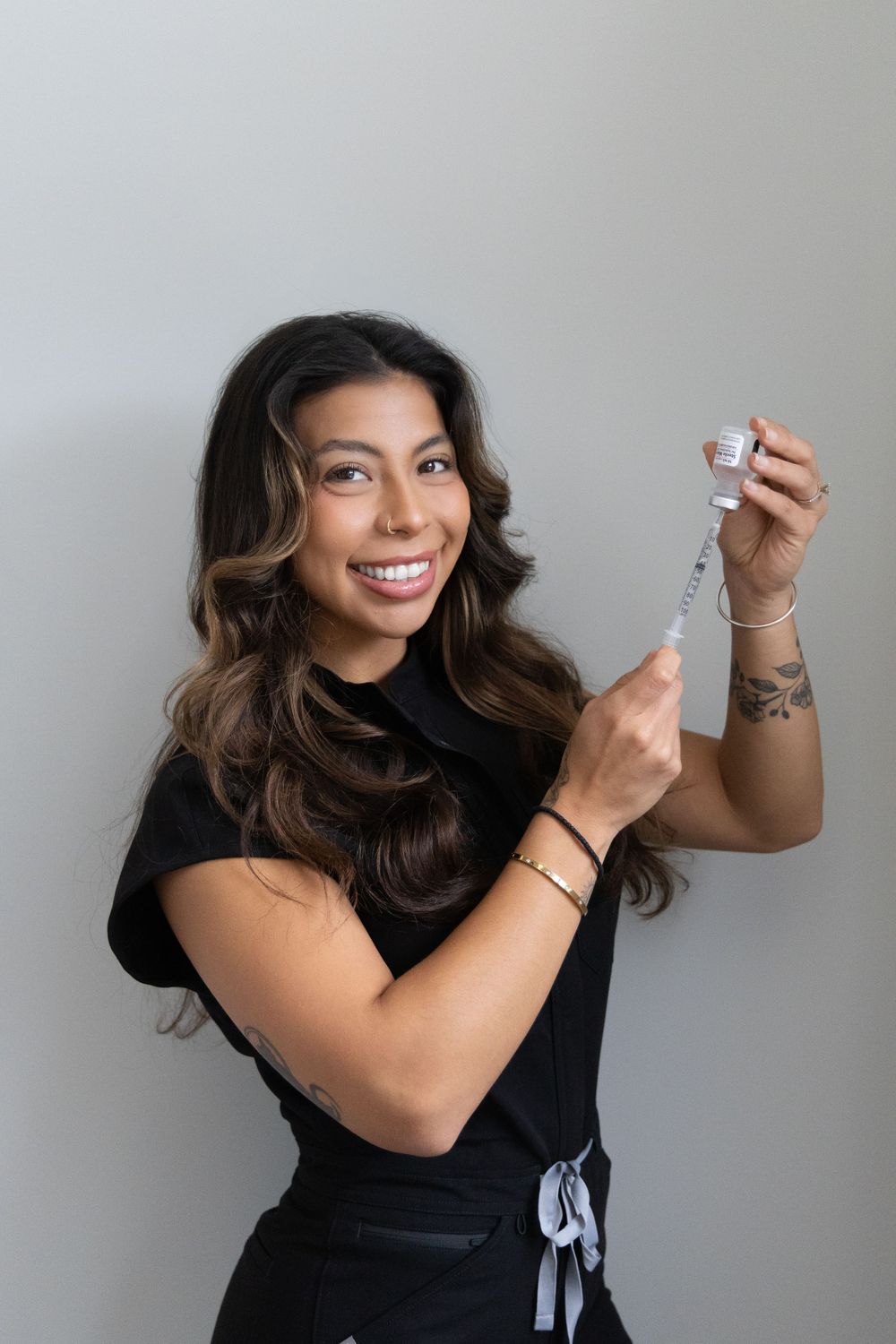 Smiling female aesthetic injector in black scrubs holding a syringe and vial for cosmetic treatment.