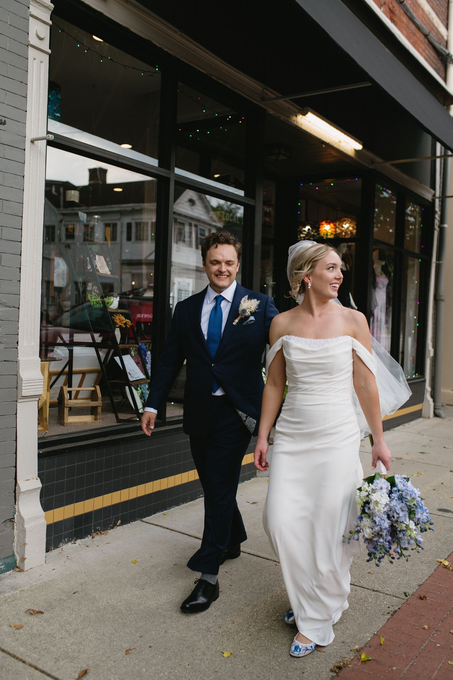 A newlywed couple walks along a storefront sidewalk with the bride holding a blue bouquet.