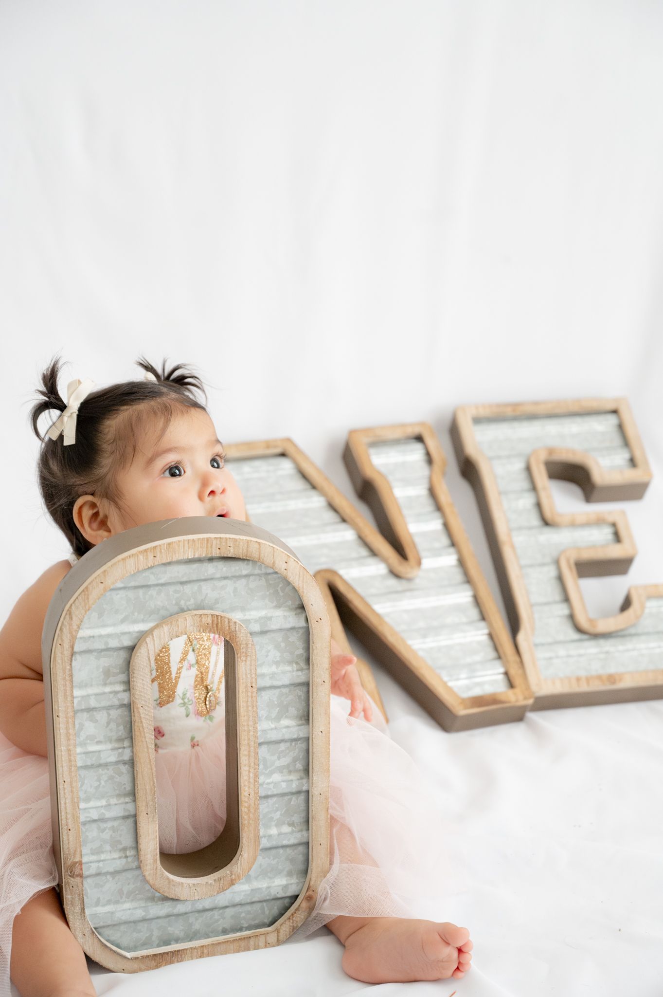 Baby in pink dress sits with decorative wooden 'ONE' letters against white backdrop.
