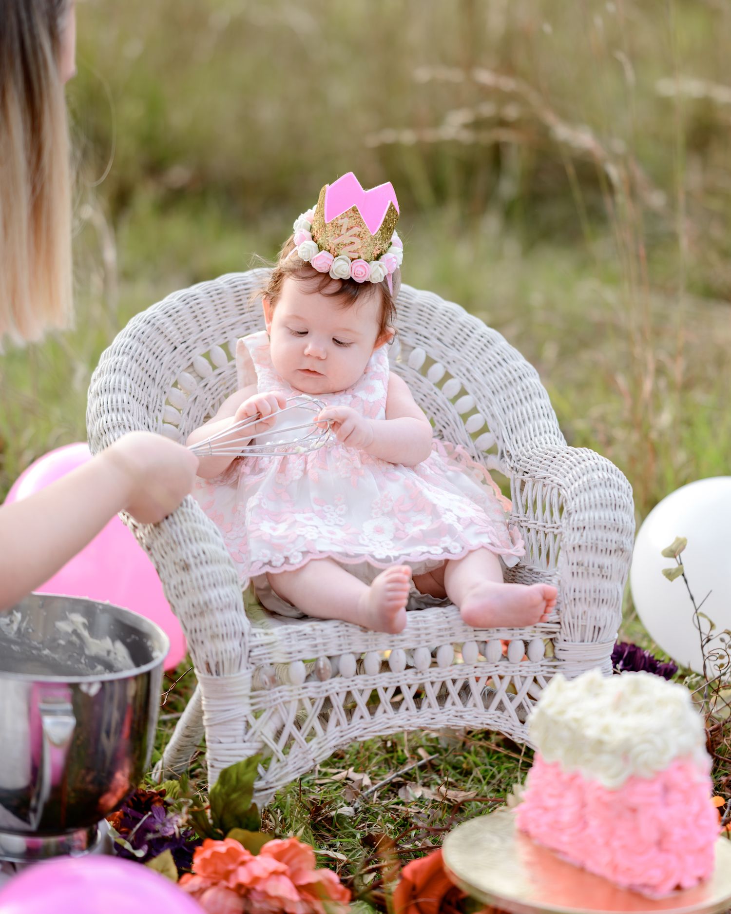Baby in pink dress sitting in white wicker chair during outdoor tea party.