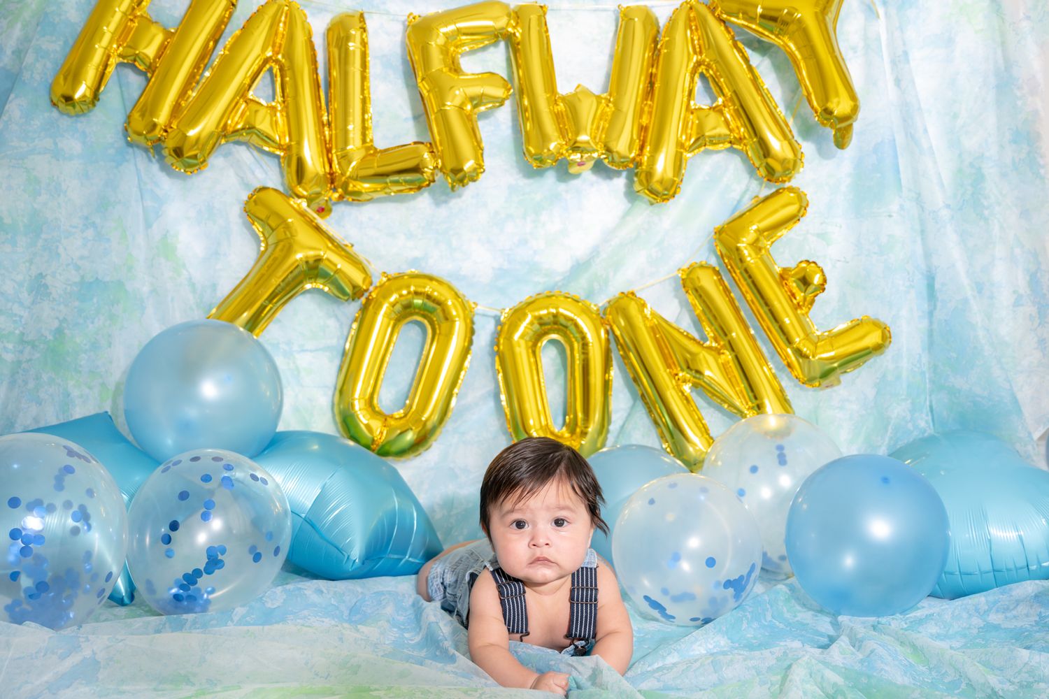 Baby lies under gold 'HALFWAY TO ONE' balloon letters with blue balloons on light background.