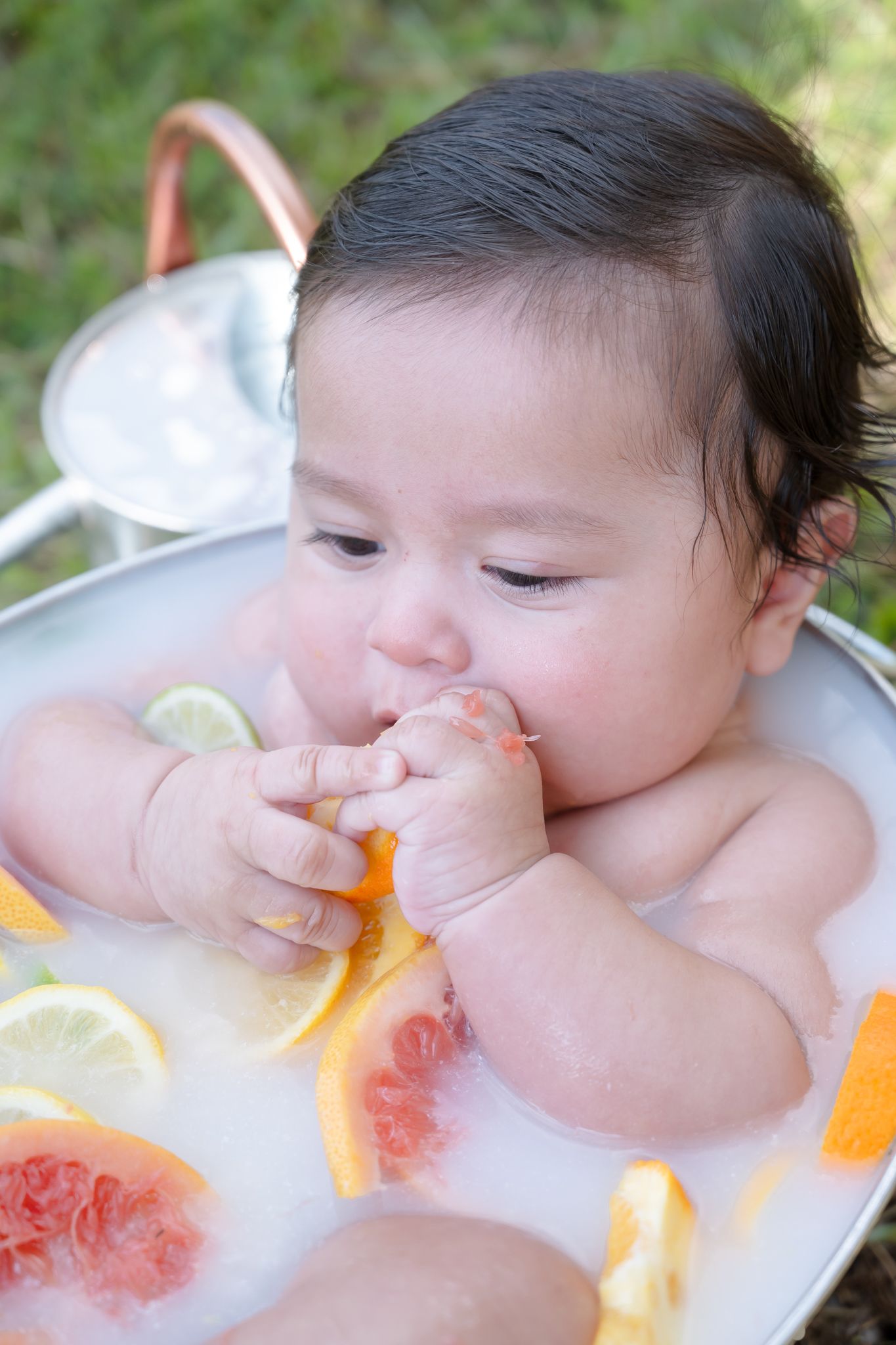 Baby enjoying a milk bath with citrus fruits and flowers in a white basin.